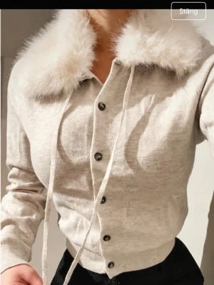 Beige kofta med fuskpäls-krage - Supersöt beige kofta med fluffig fuskpäls-krage och knappar framtill. Långa ärmar och ribbade muddar. Perfekt för dig som vill sticka ut lite extra med en trendig och mysig look.