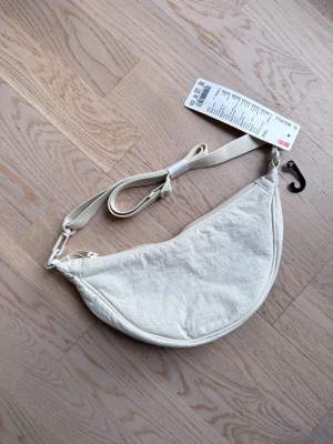 Uniqlo ultra mini bag off-white - Säljer en stilren vit axelväska från Uniqlo i halvmåneformad design. Väskan har justerbar axelrem. Perfekt för att bära dina viktigaste grejer med enkel stil.