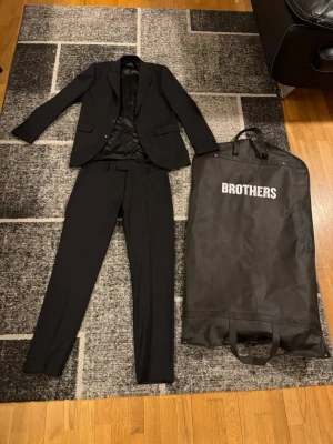 Svart kostym från Brothers - Snygg svart kostym från Brothers med klassisk kavaj och raka kostymbyxor. Kavajen har två knappar och fickor framtill, helsvart foder och stilren passform. Perfekt för dig som vill ha en tidlös och elegant look.