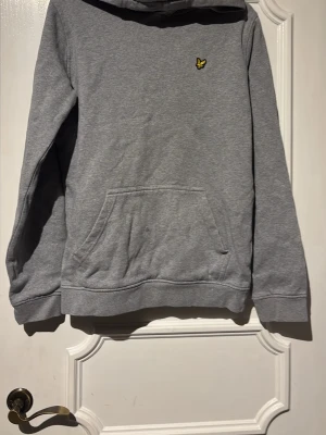 Grå hoodie från Lyle & Scott - Grå hoodie med klassisk passform från Lyle & Scott. Har en gul broderad logga på bröstet och en stor känguruficka framtill. Mjuk bomullsblandning och ribbade muddar vid ärmslut och nederkant. Perfekt för en chill och avslappnad stil.