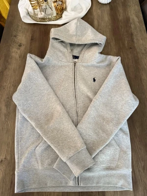 Grå hoodie från Polo Ralph Lauren - Snygg ljusgrå hoodie från Polo Ralph Lauren med dragkedja framtill och klassisk huva. Tröjan har två fickor på sidorna och den ikoniska lilla loggan broderad på bröstet. Tillverkad i mjukt bomullsmaterial som känns riktigt skönt mot huden.