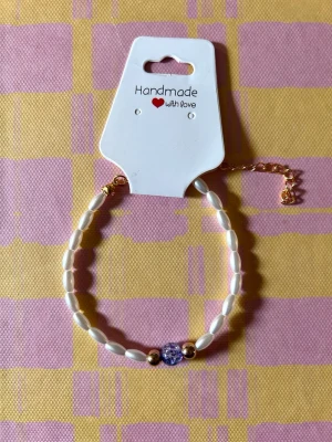 Pärlarmband med lila detalj - Snyggt handgjort armband med vita avlånga pärlor, två guldfärgade kulor och en lila glaspärla i mitten. Justerbar kedja i guldton. Perfekt accessoar för att lyfta din stil.