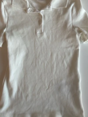 Vit piké t-shirt från Gina Tricot - Vit piké t-shirt från Gina Tricot med klassisk krage och två knappar framtill. T-shirten är kortärmad och tillverkad i mjuk bomull, perfekt för en clean och enkel stil.