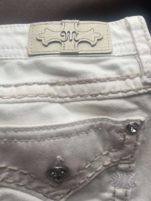 Vita miss me jeans med detaljer - Säljer ett par vita jeans från miss me med snygga kontrastsömmar och dekorativa bakfickor med metallknappar. Jeansen har en klassisk femficksmodell och tydlig logga på bakre linningen. Perfekta för dig som gillar street och premium denim.