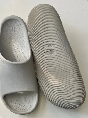 Crocs Mellow - Säljer ett par gråa Crocs mellow tofflor i mjukt och lätt gummimaterial. De har öppen tå, räfflad sula och diskret Crocs-logga på sidan. Perfekta när du vill ha något bekvämt på fötterna efter en träning eller bara att gå i. Köpta från stadium.
