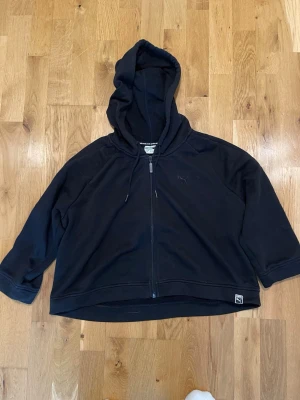 Svart hoodie med dragkedja från Puma stl S - Svart hoodie från Puma med dragkedja framtill och justerbar huva. Modellen är lite boxig och har breda ärmar. Puma-logga broderad på bröstet och liten etikett nertill. Perfekt för en chill och sportig look.