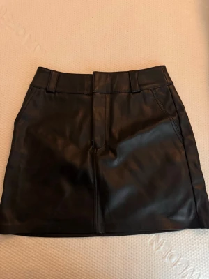 Svart skinnkjol med bälteshällor - Snygg svart kort skinnkjol med bälteshällor och klassisk design. Kjolen har en rak passform och är perfekt för att skapa en cool och trendig look. Passar till många olika stilar och är enkel att matcha med olika toppar.