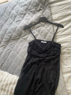 Svart volangklänning från Gina Tricot - Snygg svart klänning från Gina Tricot med smala axelband och volangdetaljer över bysten. Klänningen har en smockad överdel och är gjord i ett lätt, flowy tyg som ger en cool och trendig look.