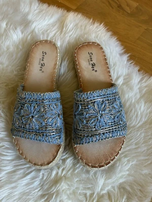 Blå broderade sandaler från Soon Her - Snygga sandaler från Soon Her med blått broderi och blommönster på ovandelen. Sandalerna har en öppen tå och flätad sula i naturmaterial som ger en bohemisk känsla. Perfekta för sommaren och lätta att matcha till olika outfits.