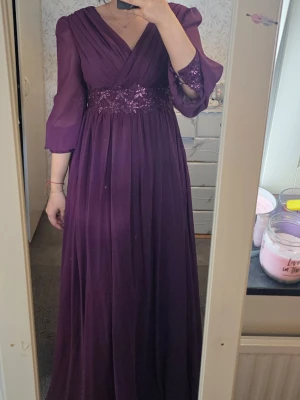 Lång lila klänning med paljettdetalj - Elegant långklänning i djup lila nyans med v-ringad omlottringning och trekvartsärmar. Midjan pryds av glittriga paljetter och spetsdetaljer som ger extra lyxig känsla. Kjolen är plisserad och faller mjukt ner till golvet. 