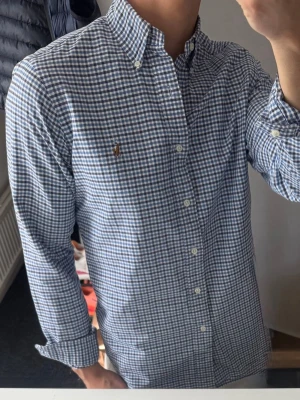 Blårutig skjorta från Ralph Lauren - Klassisk blårutig skjorta från Ralph Lauren med button-down krage och broderad logga på bröstet. Skjortan har långa ärmar, vita knappar och är tillverkad i mjuk bomull. Perfekt för en clean och stilren look. Lite missfärgad i armhålorna o kragen därav priset. Annars bra skick