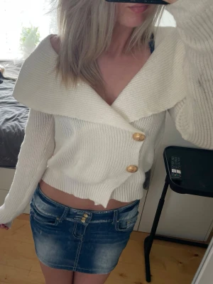Vit stickad cardigan med bred krage - Säljer en vit stickad cardigan med bred krage och två dekorativa guldknappar framtill. Tröjan har ribbstickad struktur, lång ärm och kortare passform. Perfekt för dig som vill ha en trendig och snygg look.