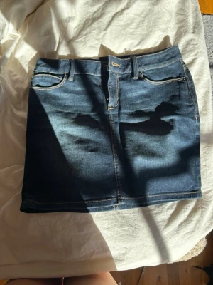 Mörkblå jeanskjol med fickor - Snygg mörkblå jeanskjol med klassisk femficksdesign och synliga sömmar. Kjolen har bälteshällor och dragkedja framtill. Perfekt för dig som gillar en enkel och tidlös stil.