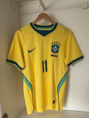 Brasilien fotbollströja Raphinha 11 - Gul Brasilien fotbollströja från Nike med gröna och blå detaljer, nummer 11 och namnet Raphinha på ryggen. Tröjan har korta ärmar, rund hals och CBF-emblem på bröstet. Perfekt för dig som älskar fotboll och vill stötta Brasilien.