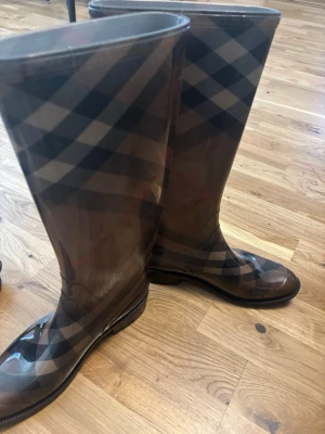 Bruna rutiga gummistövlar Burberry - Snygga bruna gummistövlar från Burberry med det klassiska rutmönstret. Höga skaft och rundad tå, perfekta för regniga dagar. Tillverkade i glansigt gummi som håller fötterna torra och ger en stilren look.