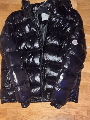 Svart glansig dunjacka från Moncler - Snygg svart dunjacka från Moncler med glansig finish och klassisk puffer-design. Jackan har huva, dragkedja framtill och Moncler-logga på ärmen. Perfekt för kalla vinterdagar och riktigt varm tack vare dunfyllningen. Går o skanna nfc