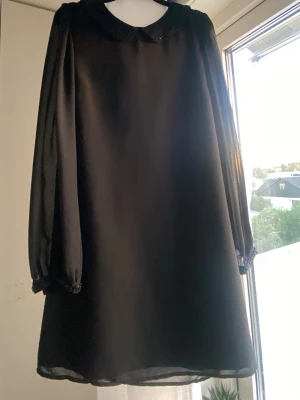 Svart kort klänning med krage - Stilren svart kort klänning med transparenta långärmar och söt krage. Klänningen har en rak passform och är tillverkad i ett lätt, flowy material som ger en luftig känsla. Med pärl detaljer på kragen och ändan av ärmen❣️