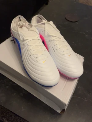 Nike Tiempo Maestro  - Säljer ett par vita Nike fotbollsskor med snygga detaljer i blått och rosa på sidorna. Skorna har klassisk snörning, svarta Nike-loggor och en modern, vågig struktur på ovandelen. Perfekta för dig som vill sticka ut på planen med både stil och funktion.