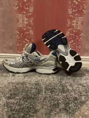 ASICS vita och blå sneakers - Snygga sneakers från ASICS i vitt och blått med silvriga detaljer. Ovandelen är i mesh och syntet för bra andningsförmåga och komfort. Klassisk sportig design med ASICS-logga på sidan och vadderad plös. Perfekta för en aktiv livsstil.