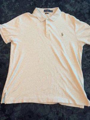Beige pikétröja från Polo Ralph Lauren - Klassisk beige pikétröja från Polo Ralph Lauren med krage, korta ärmar och knappar framtill. Tröjan har den ikoniska broderade loggan på bröstet och är tillverkad i mjuk bomull. Perfekt för en stilren och avslappnad look.