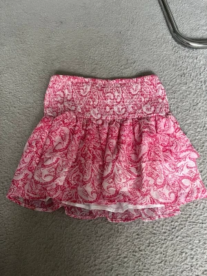 Rosa volangkjol med mönster - TRYCK GÄRNA KÖP DIRKET!!💗 Söt kortkjol i rosa och vitt med volanger och smockad midja. Kjolen har ett blommigt mönster och är tillverkad i ett lätt, luftigt material som passar perfekt till varmare dagar💗