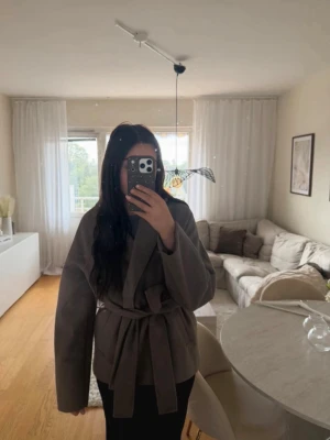 Begie/brun kappa med knytskärp - Stilren begie/brun kappa med bred krage och knytskärp i midjan. Modellen är rak och har långa ärmar, perfekt för lager-på-lager. Kappan har en minimalistisk look och passar till många olika outfits. Materialet ser ut att vara mjukt och lite tjockare för extra värme.