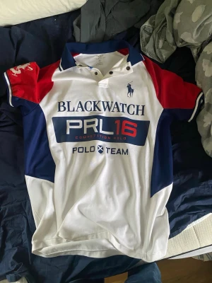 Polo Ralph Lauren Blackwatch skjorta M - Säljer en snygg Polo Ralph Lauren Blackwatch skjorta i custom fit. Skjortan är vit med blå och röda detaljer, har korta ärmar, krage och knappar vid halsen. Stora tryck på bröstet och ärmen ger en sportig vibe. Perfekt för dig som gillar klassisk men cool stil.