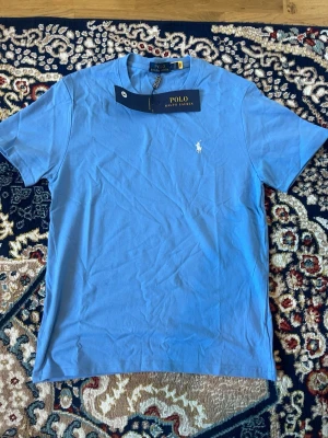 Blå t-shirt från Polo Ralph Lauren - Säljer en klassisk blå t-shirt från Polo Ralph Lauren med rund hals och det ikoniska vita logotrycket på bröstet. T-shirten är i mjuk bomull och har en normal passform. Perfekt basplagg för en clean och stilren look.