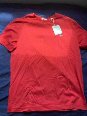 Röd t-shirt från Les Deux - Snygg röd t-shirt från gant med liten vit logga på bröstet. Klassisk passform med rund hals och korta ärmar. Tillverkad i mjuk bomull som känns skön mot huden. Perfekt för en stilren och enkel look.