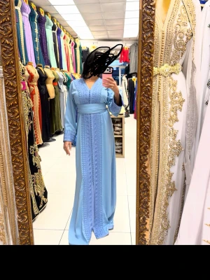 Ljusblå kaftan långklänning med broderi - Elegant ljusblå långklänning med v-ringning och broderade detaljer längs hela klänningen. Klänningen har långa ärmar och ett brett midjeband som markerar midjan. 