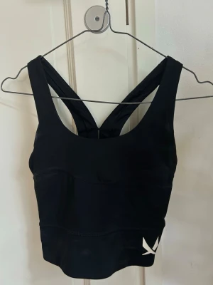 Svart croppad träningstopp - Snygg svart croppad träningstopp med breda axelband och sportig design. Toppen har en stilren look med diskret logga framtill och är tillverkad i stretchigt material som passar perfekt till träning eller en aktiv vardag.