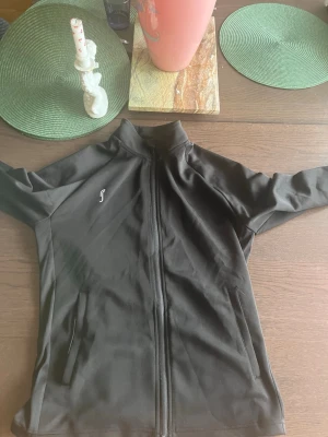 Svart träningsjacka från RS Sports - Svart träningsjacka från RS Sports med hel dragkedja framtill och två sidofickor med dragkedja. Jackan har en hög krage och är tillverkad i ett mjukt, stretchigt material som passar perfekt för träning och sportaktiviteter.