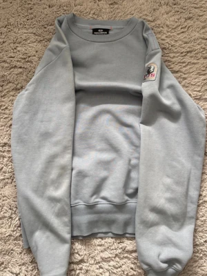 Ljusblå sweatshirt från Parajumpers - Ljusblå sweatshirt från Parajumpers med rund halsringning och ribbade muddar. Tröjan har en broderad patch på ena ärmen och är tillverkad i mjukt bomullsmaterial. Perfekt för en avslappnad och stilren look.