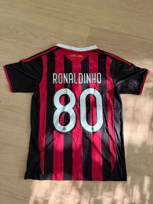 AC Milan Ronaldinho tröja Adidas - Säljer en klassisk AC Milan matchtröja med Ronaldinho och nummer 80 på ryggen. Tröjan är röd och svart med vita detaljer, har klubbmärke och Bwin-logga på bröstet samt Adidas ränder på axlarna. Perfekt för fotbollsfans som gillar retrostil.