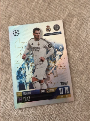Fotbollssamlarkort Brahim Díaz - Säljer ett glittrigt fotbollssamlarkort från Topps med Brahim Díaz i Real Madrid-dräkt. Kortet har detaljer i guld och visar statistik samt Champions League-märke. Perfekt för samlare av fotbollskort.