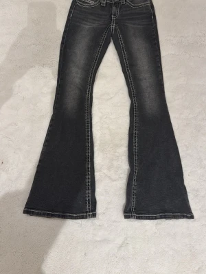 Svarta bootcut jeans med kontrastsömmar - Snygga svarta bootcut jeans med tydliga vita kontrastsömmar och slitningar framtill. Jeansen har klassisk femficksdesign och utsvängda ben som ger en retrovibe. Perfekta för dig som vill sticka ut med en cool och trendig look.