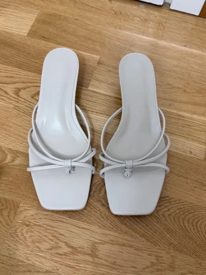 Vita pumps med smala remmar och rosett - Snygga vita pumps med smala remmar och en söt rosettdetalj framtill. Skorna har fyrkantig tå och låg klack, vilket ger en trendig och stilren look. Tillverkade i ett glansigt material som ger en fräsch känsla.