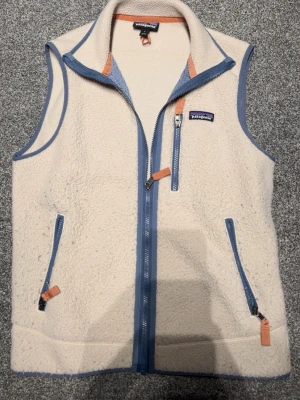 Beige fleeceväst från Patagonia - Snygg beige fleeceväst från Patagonia med blå detaljer och dragkedjor. Västen har tre fickor med dragkedja och orangea dragflikar. Perfekt för lager-på-lager och outdoor-vibe. Mjuk och varm med hög krage och klassisk Patagonia-logga på bröstet.