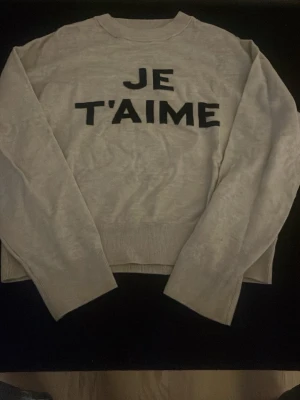Beige tröja med texten JE T'AIME - Snygg beige tröja med svart text 'JE T'AIME' framtill. Tröjan har rund halsringning, långa ärmar och ribbade muddar. Perfekt för dig som gillar statement-plagg och vill ha en enkel men trendig look.