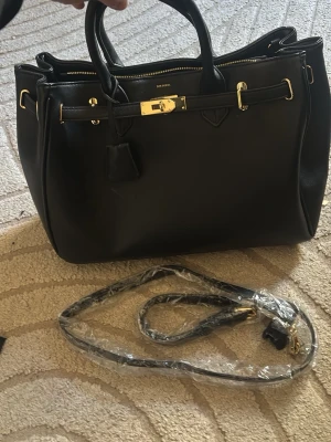 Svart handväska med gulddetaljer - Stilren svart handväska med två rejäla handtag och snygga guldfärgade detaljer. Väskan har en klassisk boxig form och är tillverkad i slätt konstläder. Perfekt för dig som gillar en elegant look och vill ha plats för allt du behöver. H liknande väska🩷 Köpt ca 1500kr