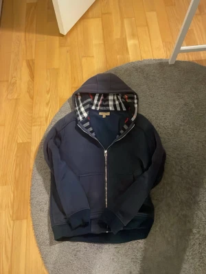 Marinblå hoodie från Burberry - Snygg marinblå hoodie från Burberry med dragkedja och klassiskt rutigt foder i hu for extra stilpoäng. Huva med dragsko och två fickor framtill. Perfekt för dig som vill ha en exklusiv och bekväm look. Materialet känns mjukt och kvalitativt. Dragtjedjehållaren har gått sönder har behövt bytt den med en reserv del