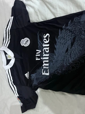 Svart Real Madrid tröja Adidas - Cool svart Real Madrid matchtröja från Adidas med vitt tryck och detaljer. Tröjan har ett drakmönster på framsidan, vit krage och vita ränder på axlarna. Fly Emirates-logga på bröstet och Ronaldo 7 på ryggen. Perfekt för fotbollsfans!