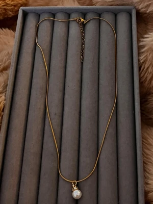 Guldhalsband med pärlhänge - Snyggt halsband i guldfärgad metall med ett elegant vitt pärlhänge. Kedjan är tunn och smidig med justerbar längd. Perfekt för dig som gillar stilrena accessoarer med en klassisk touch.