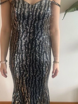 Glittrig långklänning med silvermönster - Säljer en elegant långklänning med glittriga silverpaljetter och svarta inslag. Klänningen har smala axelband och en v-ringad front. Perfekt för dig som vill sticka ut och glänsa på balen eller festen. Materialet är polyester och klänningen har en tight passning.den passar 38,40,42 stretchigt material.