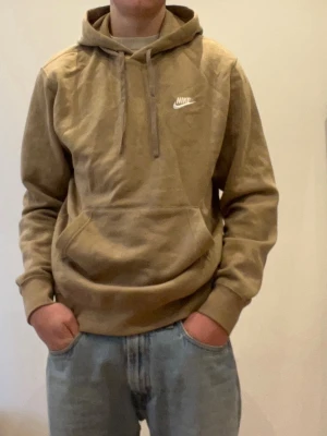 Beige hoodie från Nike med snörning - Säljer en beige hoodie från Nike med klassisk logga på bröstet. Hoodien har snörning i huvan, ribbade muddar och en stor magficka. Perfekt för dig som gillar en avslappnad och stilren look.