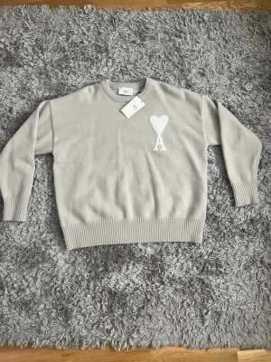 Ami Paris Beige stickad tröja  - Snygg beige stickad tröja från Ami Paris med rund halsringning och ribbade muddar. Tröjan har Ami:s ikoniska hjärta och A-logga broderad i vitt på bröstet. Perfekt för dig som gillar stilrena och trendiga plagg.