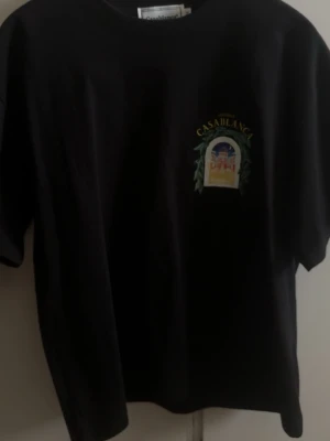 Svart Casablanca t-shirt med tryck - Säljer en svart t-shirt från Casablanca med färgglatt tryck av ett palats och gröna blad både fram och bak. T-shirten har rund hals, korta ärmar och en relaxed passform. Perfekt för dig som vill ha något unikt och stilrent i garderoben.