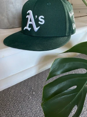 Grön Oakland A's keps från New Era *Nyskick - Snygg grön keps med Oakland A's logga i vitt framtill och broderad text på sidan. Klassisk flat brim och New Era-märke. Perfekt för dig som gillar sportiga accessoarer och baseballstil. I nyskick!! 