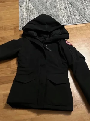 Svart dunjacka från Canada Goose - Säljer en svart dunjacka från Canada Goose med huva och klassisk logga på ärmen. Jackan har dragkedja, två stora fickor framtill och en ficka på ärmen. Behöver pengar snabbt så de därför den säljs så billigt 