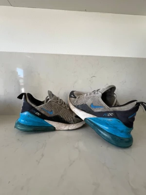 Nike Air Max 270 grå/blå sneakers - Nike Air Max 270 sneakers i grå mesh med svarta och blå detaljer samt stor blå Air-sula. Skorna har en sportig look med ventilerande material och tydlig logga på sidan. Perfekta för dig som gillar streetwear och vill ha något bekvämt och snyggt.